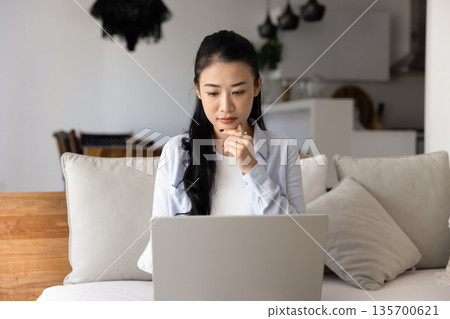 Pensive Asian woman sits on sofa with laptop 135700621