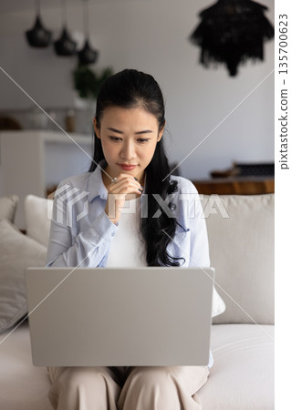 Engrossed Vietnamese woman working on laptop seated on couch Engrossed Vietnamese woman working on laptop seated on couch 135700623