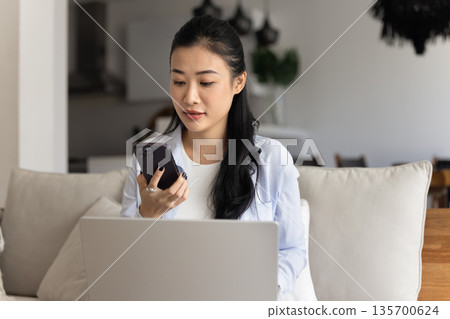 Asian woman sits on sofa with laptop and using smartphone Asian woman sits on sofa with laptop and using smartphone 135700624