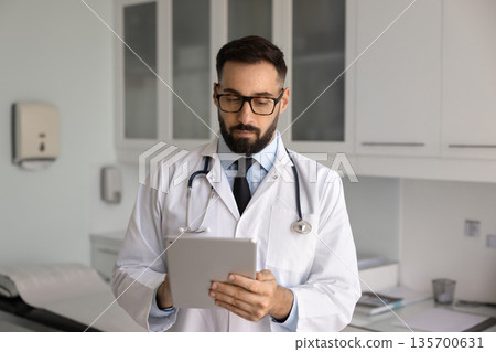 Serious busy Latin doc in stylish eyeglasses typing on tablet Serious busy Latin doc in stylish eyeglasses typing on tablet 135700631
