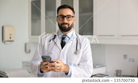 Positive young Hispanic physician man in lab coat holding smartphone Positive young Hispanic physician man in lab coat holding smartphone 135700730