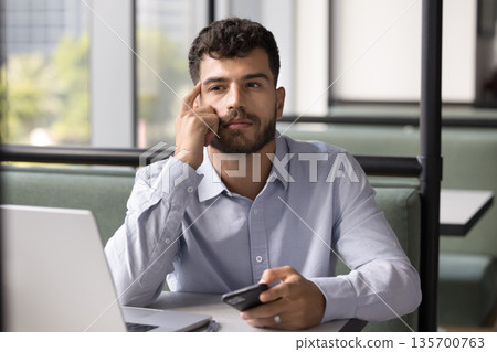 Pensive young man sitting at table contemplating message on cell Pensive young man sitting at table contemplating message on cell 135700763