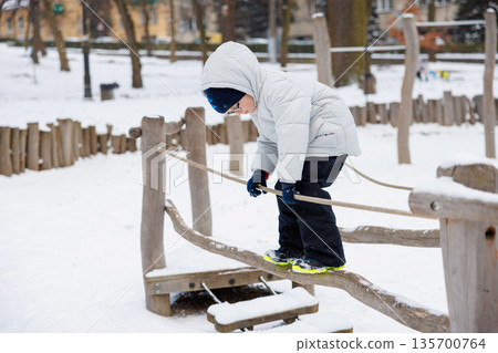 Child Balancing on Rope Bridge in Snowy Playground Child Balancing on Rope Bridge in Snowy Playground 135700764
