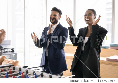 Laughing businesspeople standing by kicker table greeting colleagues with winning Laughing businesspeople standing by kicker table greeting colleagues with winning 135700862