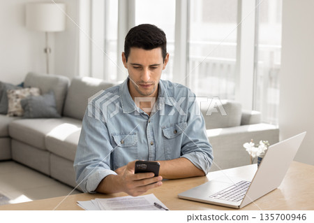 Focused young male remote working professional multitasking using phone notebook Focused young male remote working professional multitasking using phone notebook 135700946