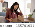 Woman sit on sofa opens cardboard package and feels upset 135701031