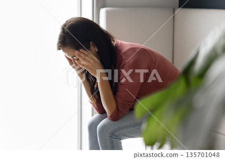 Sad woman covering face in despair seated indoors Sad woman covering face in despair seated indoors 135701048