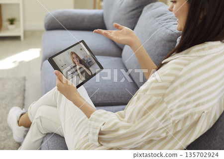 Young woman relaxing on cozy sofa with tablet and talking with friend using video call app 135702924