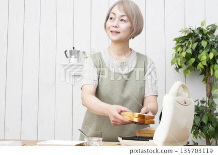 一位老婦人正在用熱三明治機製作熱三明治。 一位老婦人正在用熱三明治機製作熱三明治。 135703119