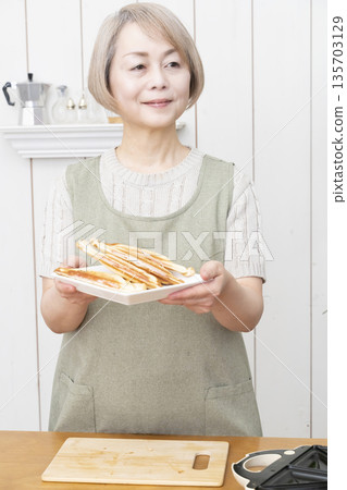 一位老婦人正在用熱三明治機製作熱三明治。 一位老婦人正在用熱三明治機製作熱三明治。 135703129