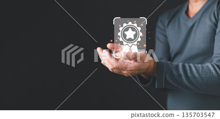 A person in a gray sweater holds a glowing digital icon of a certificate badge with a star, symbolizing quality, trust, and achievement in modern business standards and recognition. A person in a gray sweater holds a glowing digital icon of a certificate badge with a star, symbolizing quality, trust, and achievement in modern business standards and recognition. 135703547