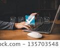 A person using a laptop and smartphone interacts with glowing digital icons of documents and a large checkmark, symbolizing document approval, verification, and secure online processing. 135703548