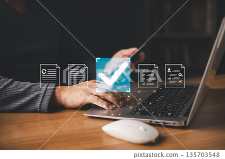 A person using a laptop and smartphone interacts with glowing digital icons of documents and a large checkmark, symbolizing document approval, verification, and secure online processing. 135703548