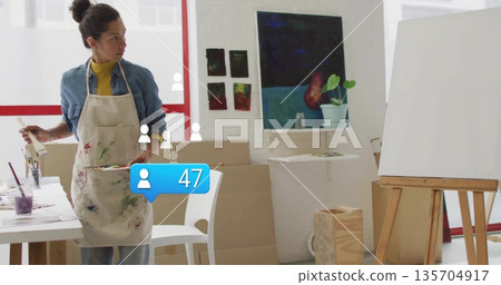 Mature artist woman mixing paint on palette in creative studio, with easel and canvases 135704917