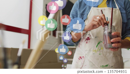 Painter wearing apron mixing brushes in jar at studio, with floating thumbs-up icons, copy space Painter wearing apron mixing brushes in jar at studio, with floating thumbs-up icons, copy space 135704931