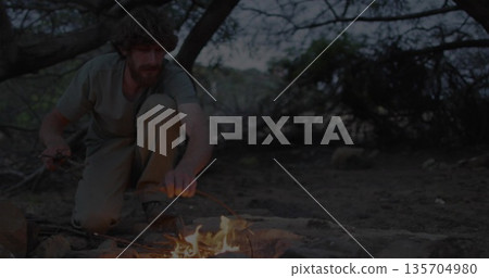 Kneeling man in cargo pants arranging sticks in stone ring in forest clearing, with campfire glow 135704980