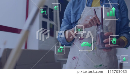 Artist stirring paint in glass jar with brush in studio, wearing stained apron, with overlay icons 135705083