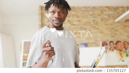 Artist with dreadlocks in paint-stained shirt pointing brush toward camera in studio, with canvases Artist with dreadlocks in paint-stained shirt pointing brush toward camera in studio, with canvases 135705127