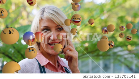 Smiling woman wearing green apron taking call on smartphone in garden, with floating emoji spheres Smiling woman wearing green apron taking call on smartphone in garden, with floating emoji spheres 135705141