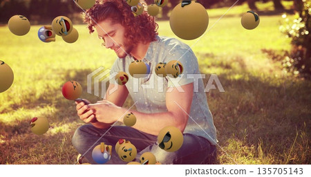 Smiling adult male holding smartphone while sitting on park lawn, with floating emoji spheres Smiling adult male holding smartphone while sitting on park lawn, with floating emoji spheres 135705143