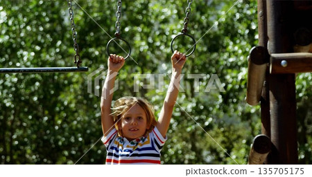 Boy wearing striped shirt hanging from rings in playground under sunlit green trees, chains beams Boy wearing striped shirt hanging from rings in playground under sunlit green trees, chains beams 135705175