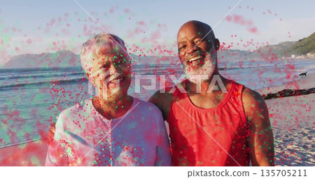 Standing couple wearing red tank top and light shirt on sandy beach, with colored powder drifting 135705211