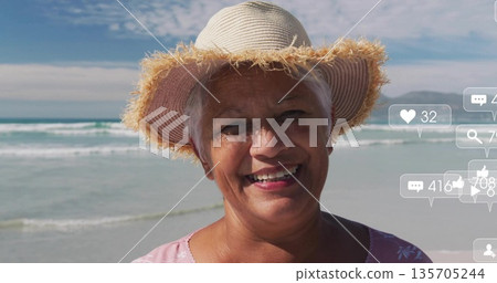 Smiling senior woman wearing straw sun hat and pink floral top at beach, floating reaction icons Smiling senior woman wearing straw sun hat and pink floral top at beach, floating reaction icons 135705244