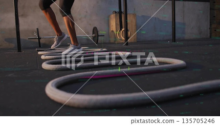 Stepping woman in black leggings and trainers on gym mat, battle rope loops, barbell, copy space Stepping woman in black leggings and trainers on gym mat, battle rope loops, barbell, copy space 135705246