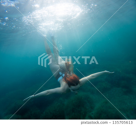 Underwater woman diving through clear turquoise ocean water as sunbeams shine from the surface promoting freedom and adventure in nature 135705613