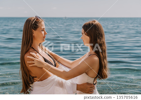 Women beach hugging as they look at each other while standing in the calm blue sea water on a bright summer day Women beach hugging as they look at each other while standing in the calm blue sea water on a bright summer day 135705616