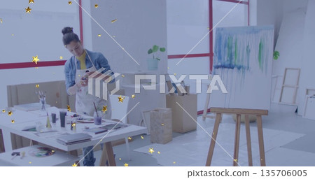 Holding smartphone, female artist in paint apron examining canvas drips at art studio, with easel 135706005