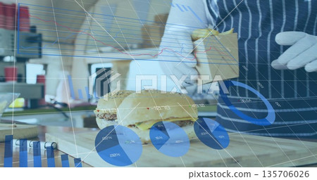Cook wearing striped apron with gloves lifting burger into box at prep station with data overlay 135706026