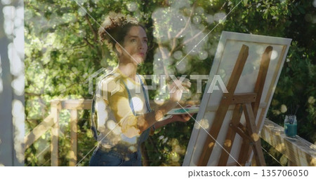 Painting Hispanic woman wearing overalls and holding paintbrush before canvas on deck with easel Painting Hispanic woman wearing overalls and holding paintbrush before canvas on deck with easel 135706050
