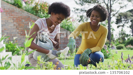 Kneeling mother and daughter pouring water on flowering plants in backyard, with gray watering can Kneeling mother and daughter pouring water on flowering plants in backyard, with gray watering can 135706102