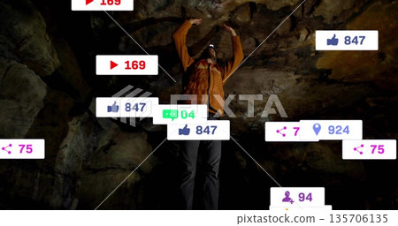Reaching explorer in orange jacket with headlamp touching cave ceiling amid social media overlays 135706135