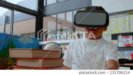 Exploring virtual reality world boy wearing VR headset at classroom table, with globe and books 135706214