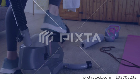 Pedaling woman in dark leggings on stationary bike in home gym, with foam roller and kettlebell Pedaling woman in dark leggings on stationary bike in home gym, with foam roller and kettlebell 135706232