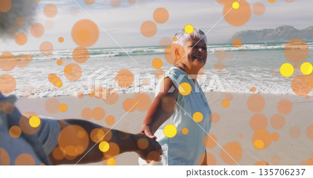 Walking senior woman holding hand of man along sandy shoreline, wearing sleeveless blouse Walking senior woman holding hand of man along sandy shoreline, wearing sleeveless blouse 135706237
