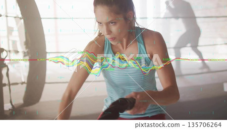 Woman in teal top, red leggings gripping battle ropes in gym with punching bag, digital overlay 135706264