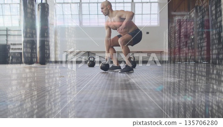 Crouching shirtless man lifting kettlebells in gym studio, with hanging punching bags and bench Crouching shirtless man lifting kettlebells in gym studio, with hanging punching bags and bench 135706280