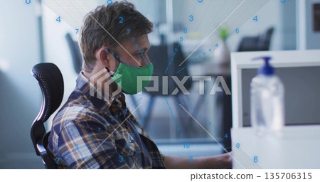 Man with green mask analyzing numeric data on computer monitor at desk with sanitizer, copy space 135706315
