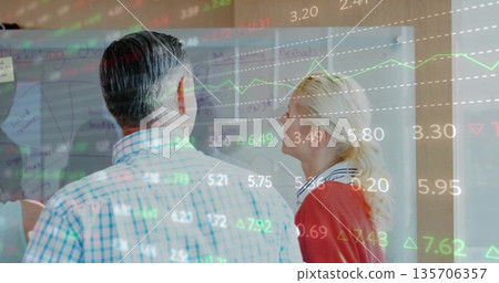 Analyzing coworkers studying data projection on transparent board in office, with sticky notes Analyzing coworkers studying data projection on transparent board in office, with sticky notes 135706357