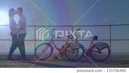 Kissing couple standing on paved waterfront walkway at sunset, with rental bicycles, copy space 135706400
