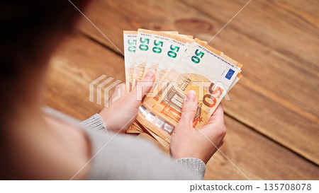 Over the shoulder view of a woman counting euro banknotes, focusing on hands and money, symbolizing personal finance, savings, and budgeting. 135708078