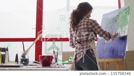 Hispanic artist wearing apron painting on wooden easel canvas in studio, with floating AR icons 135708346