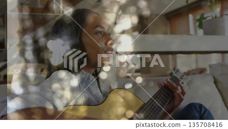 Strumming acoustic guitar musician wearing denim outfit on couch in living room, with bokeh lights Strumming acoustic guitar musician wearing denim outfit on couch in living room, with bokeh lights 135708416