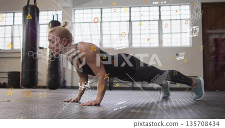 Female athlete wearing sportswear performing plank push-up on gym mat, with punching bags Female athlete wearing sportswear performing plank push-up on gym mat, with punching bags 135708434