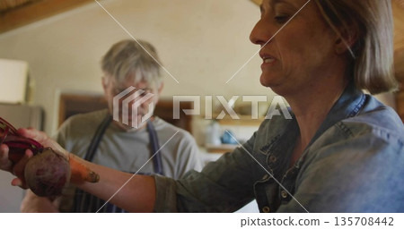 Denim-shirt woman handing fresh beets over wooden countertop to apron-clad man in farmhouse kitchen Denim-shirt woman handing fresh beets over wooden countertop to apron-clad man in farmhouse kitchen 135708442