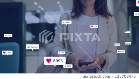 Standing businesswoman in blazer holding smartphone and handbag in lobby, showing reaction icons 135708478