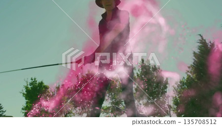 Swinging senior woman holding golf club on golf course, wearing sun hat with swirling pink powder 135708512